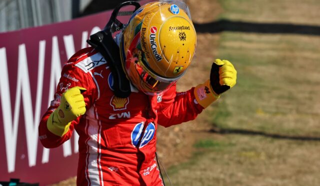Race car driver in a red suit with yellow gloves celebrates with fists raised near a track barrier.