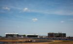 Race car speeding on a curved track with grandstands, banners, and a blue sky in the background.