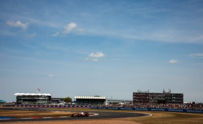 Race car speeding on a curved track with grandstands, banners, and a blue sky in the background.