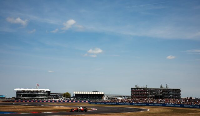 Race car speeding on a curved track with grandstands, banners, and a blue sky in the background.