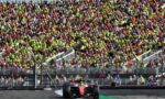 Red Formula 1 car on a race track with a packed grandstand behind barriers and spectators cheering nearby.”,