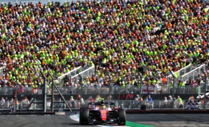 Red Formula 1 car on a race track with a packed grandstand behind barriers and spectators cheering nearby.”,