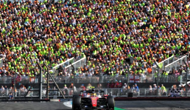Red Formula 1 car on a race track with a packed grandstand behind barriers and spectators cheering nearby.”,