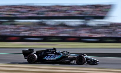 Black Mercedes Formula 1 car speeding on a racetrack with a blurred grandstand audience in the background, sponsor logos visible on the car.