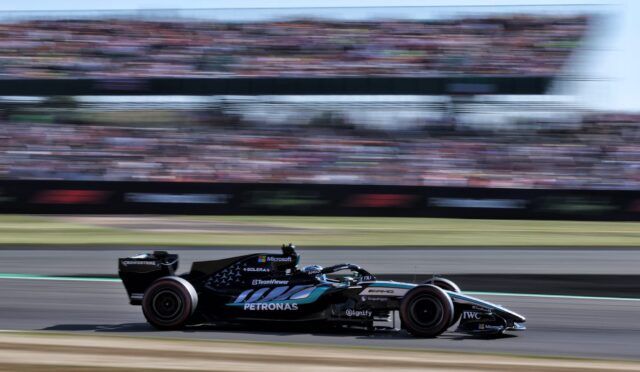 Black Mercedes Formula 1 car speeding on a racetrack with a blurred grandstand audience in the background, sponsor logos visible on the car.