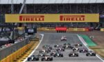 Multiple Formula 1 cars race down a track at the start, beneath large Pirelli banners in a crowded grandstand.