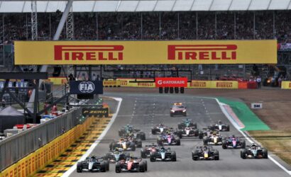 Multiple Formula 1 cars race down a track at the start, beneath large Pirelli banners in a crowded grandstand.