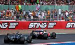 Formula racing cars speed around a circuit as a packed grandstand watches behind a safety fence, with Sting Energy Drink banners along the barrier.