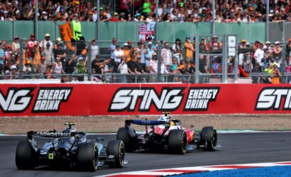 Formula racing cars speed around a circuit as a packed grandstand watches behind a safety fence, with Sting Energy Drink banners along the barrier.