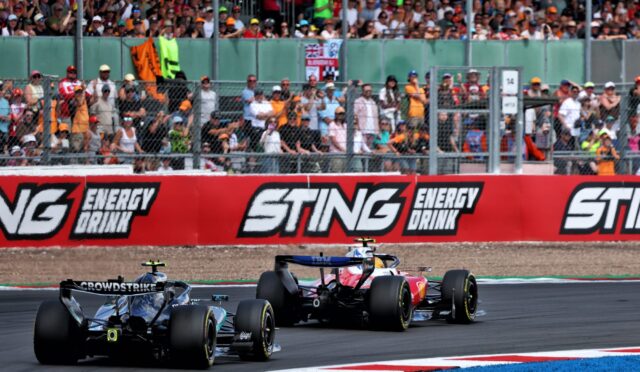 Formula racing cars speed around a circuit as a packed grandstand watches behind a safety fence, with Sting Energy Drink banners along the barrier.