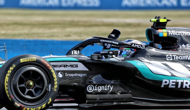 Close-up of a Formula 1 car's side with sponsor logos (Microsoft, AMD) and a Pirelli tire on a race track barrier in the background.