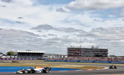 Formula 1 cars race along a curved track with a large multi-level grandstand full of spectators in the background.