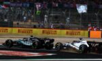 Two Formula 1 cars race side by side on a track, with sponsor boards and a cheering crowd in the stands behind them.