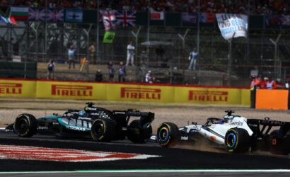 Two Formula 1 cars race side by side on a track, with sponsor boards and a cheering crowd in the stands behind them.