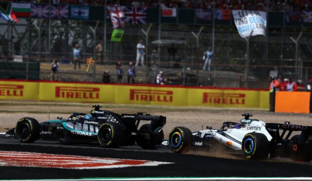 Two Formula 1 cars race side by side on a track, with sponsor boards and a cheering crowd in the stands behind them.