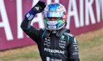 Race car driver in a dark racing suit and colorful helmet waves to the crowd, sponsor logos visible on the suit.