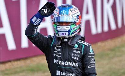 Race car driver in a dark racing suit and colorful helmet waves to the crowd, sponsor logos visible on the suit.
