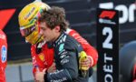 Two team members share a hug on a racing pit area, one in a red suit with a yellow-helmeted driver and sponsor logos visible in the background.