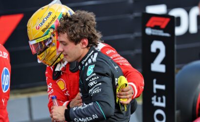 Two team members share a hug on a racing pit area, one in a red suit with a yellow-helmeted driver and sponsor logos visible in the background.