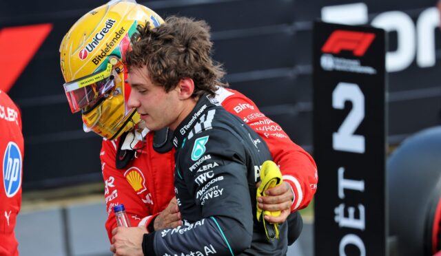 Two team members share a hug on a racing pit area, one in a red suit with a yellow-helmeted driver and sponsor logos visible in the background.