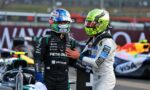 Two race car drivers in helmets stand close, one in black Petronas suit and the other in white, in a pit area with a race car in the background.