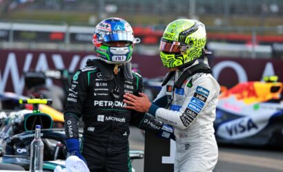 Two race car drivers in helmets stand close, one in black Petronas suit and the other in white, in a pit area with a race car in the background.