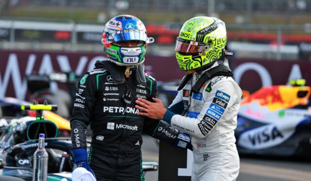 Two race car drivers in helmets stand close, one in black Petronas suit and the other in white, in a pit area with a race car in the background.