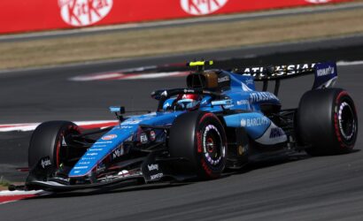 Two blue Formula 1 cars race side by side on a track, sponsor logos visible on the cars and red advertising barriers in the background.