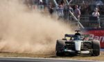 Mercedes F1 car speeds through a dusty corner, kicking up a large cloud of dirt as spectators watch from behind a chain-link fence.