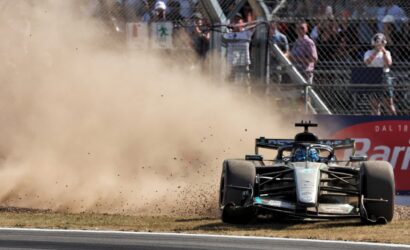 Mercedes F1 car speeds through a dusty corner, kicking up a large cloud of dirt as spectators watch from behind a chain-link fence.