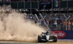 Formula 1 car on a dusty track kicking up a wide dust cloud; stands and banners in the background.