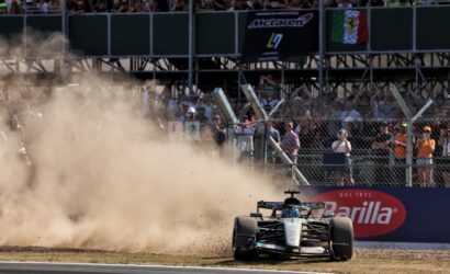 Formula 1 car on a dusty track kicking up a wide dust cloud; stands and banners in the background.