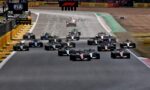 Formula 1 cars line up at the start on a race track, with sponsor banners overhead and barriers along the sides.