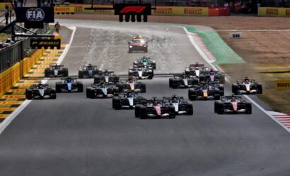 Formula 1 cars line up at the start on a race track, with sponsor banners overhead and barriers along the sides.