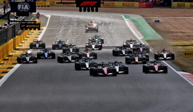 Formula 1 cars line up at the start on a race track, with sponsor banners overhead and barriers along the sides.