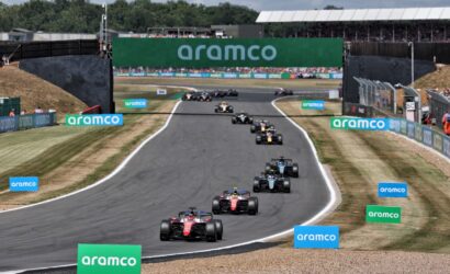 A line of Formula 1 cars race down a winding track, with green Aramco banners along the barriers and a large Aramco sign spanning a bridge above the course.
