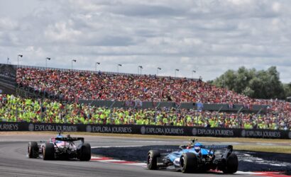 Formula 1 race cars on a track, a packed grandstand of spectators under a cloudy sky, with Explore Journeys banners along the barrier.