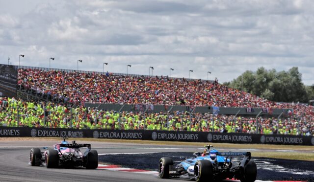 Formula 1 race cars on a track, a packed grandstand of spectators under a cloudy sky, with Explore Journeys banners along the barrier.