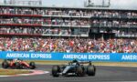 Formula One cars race along a track with a packed grandstand behind, blue American Express banners along the barrier.