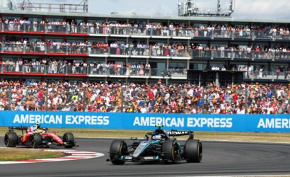 Formula One cars race along a track with a packed grandstand behind, blue American Express banners along the barrier.