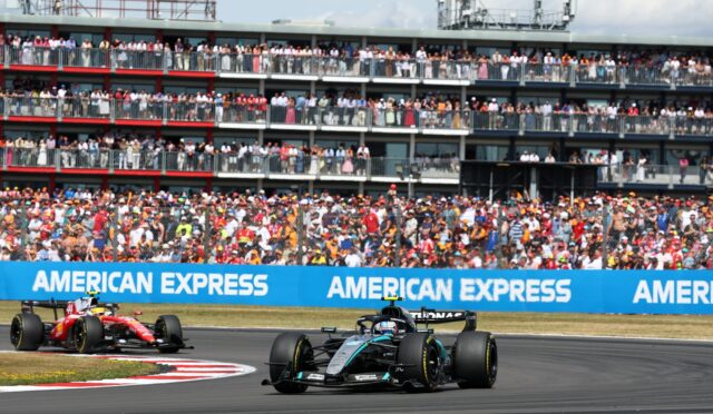 Formula One cars race along a track with a packed grandstand behind, blue American Express banners along the barrier.