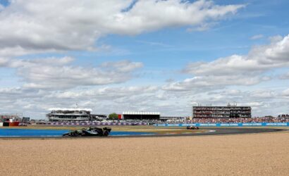 Black Formula 1 car speeding along a track with gravel in the foreground and grandstands full of spectators in the distance under a partly cloudy sky.