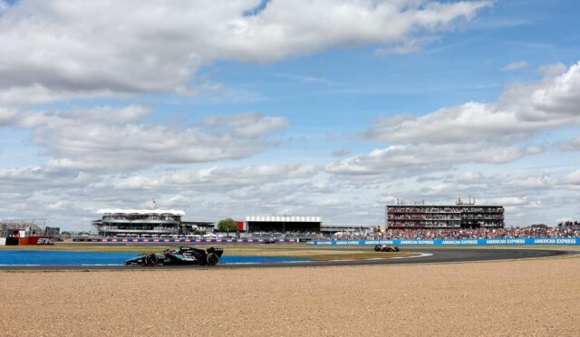 Black Formula 1 car speeding along a track with gravel in the foreground and grandstands full of spectators in the distance under a partly cloudy sky.