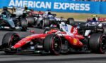 Red Ferrari Formula 1 car #16 leads a pack on a grey race track, with other F1 cars closely behind and a blue American Express barrier in the background.