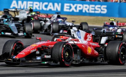 Red Ferrari Formula 1 car #16 leads a pack on a grey race track, with other F1 cars closely behind and a blue American Express barrier in the background.