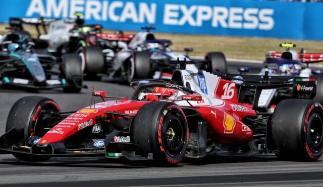 Red Ferrari Formula 1 car #16 leads a pack on a grey race track, with other F1 cars closely behind and a blue American Express barrier in the background.