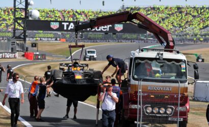 Red recovery truck with a crane hoists a race car off the track as a crowded grandstand watches in the background.