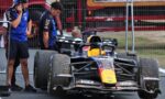 Formula 1 car in the pit area with two crew members standing beside, one on a phone, behind a metal fence.