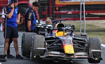 Formula 1 car in the pit area with two crew members standing beside, one on a phone, behind a metal fence.