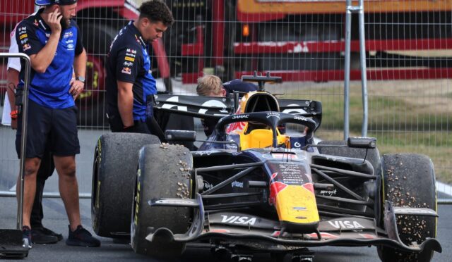 Formula 1 car in the pit area with two crew members standing beside, one on a phone, behind a metal fence.